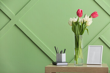 Vase with tulips, stationery supplies and blank photo frame on table near color wall