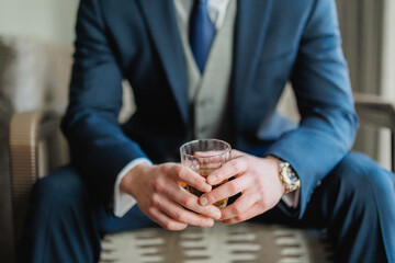 The groom in a blue suit and blue tie, the businessman sits and holds a glass of whiskey Preparing the groom for the wedding morning Close-up Cropped photo