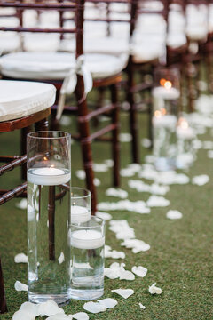 A glass vases with burning candles inside and white rose petals decoration Cropped photo Close up Lit candles in transparent glass jars on the ground near the row of wooden chairs 