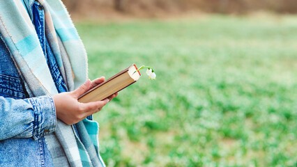 Reading book concept. Brunette woman in warm sweater and denim jacket at nature. Hipster young girl on a walk in spring.