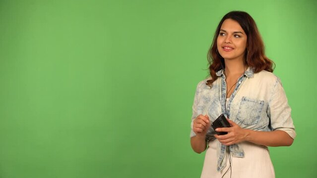 A young beautiful Caucasian woman listens to music on a smartphone with earphones on and dances with a smile - green screen background
