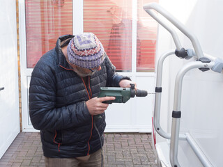 A man drills into his motorhome rear bike rack to make a hole for securing with a bolt.He has a corded power drill.Wears a hat and glasses