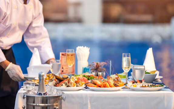 Waiter in white shirt uniform serving dishes of seafood, king pron, crab, champagne, wine on an outdoor table at wedding party or hotel yard garden event. Dinner food catering idea. Selective focus