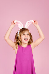 Easter concept - happy little caucasian girl raise head up touching bunny ears headband over pink background and looking away, copy space , vertical orientation