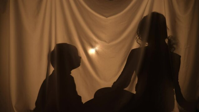 Silhouette of woman giving boy gift in form of toy house. Mom and little son spend leisure together, sitting behind curtain at home. Puppet theater of shadows, play of shadow and light. Close up.