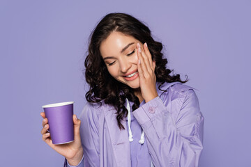 joyful young woman in raincoat holding paper cup isolated on purple.