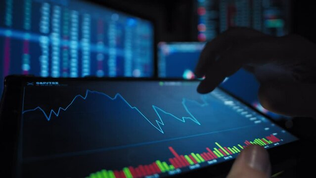 A trader holds a tablet in his hands looks and analyzes stock market graphics against the defocused background with market date close-up slow motion