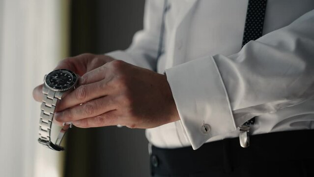 Men in a white shirt and black straps putting on his silver luxury watch in hotel room looking very elegant - close up slowmotion