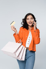 amazed woman talking on smartphone while holding credit card with cashback and shopping bags isolated on grey.