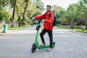 Hipster man with electric scooter outdoors in park. Young attractive student with backpack is...