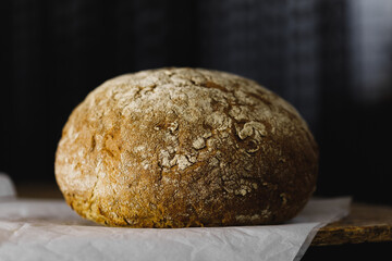 round bread with a bright crispy crust lies on parchment on a wooden table, dark background, side view