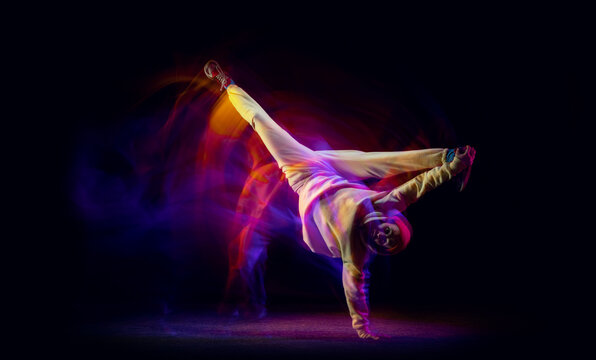 Solo dance. Young flexible sportive man dancing hip-hop or breakdance in white outfit on dark background in mixed yellow neon light. Beauty, sport, youth, action, moves