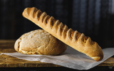 round bread and baguette bread with a bright crispy crust lies on parchment on a wooden table, dark background, side view