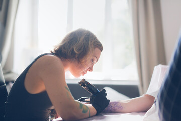 Tattoo parlor, pretty European red-haired girl makes a tattoo on arm. Woman creates a tattoo on a...