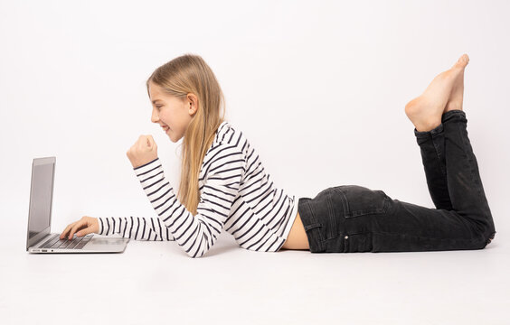 Side view of smiling young girl in striped t-shirt and jeans lying on the floor and using laptop computer over white background