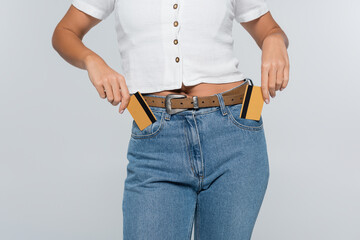 cropped view of young woman putting credit cards with cashback in pockets isolated on grey.