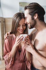 happy man hugging smiling girlfriend in pink silk robe holding cup of coffee.