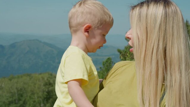 Pretty mother with long loose hair cuddles chubby cheeks little son against old distant mountains in mist at resort closeup slow motion