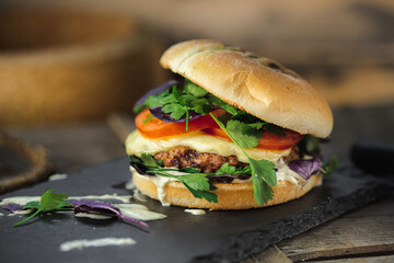 burger with beef patty, melted cheese, cream sauce, tomatoes, garnished with parsley and basil, stands on a stone blue stand, on a wooden surface, close-up