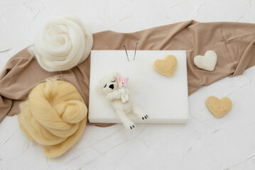 A small sleeping woolen white teddy bear made by dry felting. Felt toy for a newborn photo session. handmade product. Workplace for dry felting with a foam sponge, needles, fabric and skeins of wool 