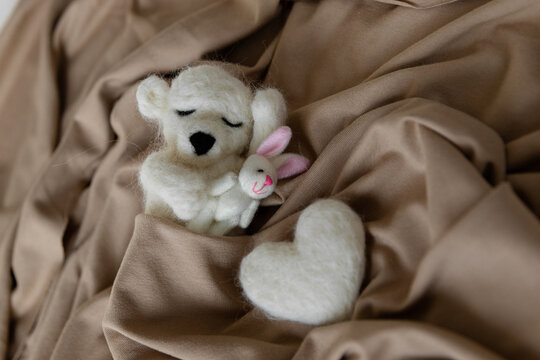 A small sleeping woolen white bear cub hugging a tiny hare and a heart made of wool lies nearby. Toys made by dry felting. Felt toy for a newborn photo session. Handmade product