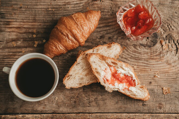 cut croissant with butter and strawberry jam with a cup of coffee, on a wooden background, top view.