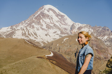 Naklejka na ściany i meble A young beautiful girl in a blue long dress and a denim jacket is sitting against the backdrop of the high Kazbek mountains in the rays of the dawn sun. Georgia. Europe