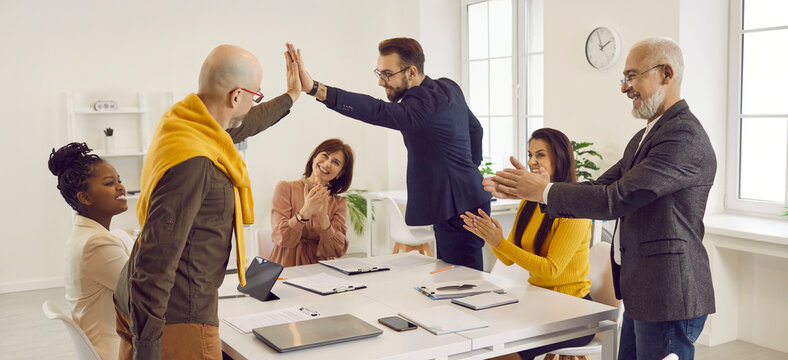 Two young men high five each other while multiracial colleagues are applauding. Team of happy motivated business people celebrating results of successful teamwork during work meeting in boardroom
