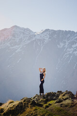 Obraz premium A beautiful girl in a long dress stands posing against the backdrop of the high mountains in the rays of the dawn sun. Europe