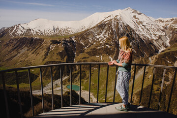 Fototapeta na wymiar A girl with long hair stands against the backdrop of a mountain landscape with snow-capped peaks and a beautiful lake