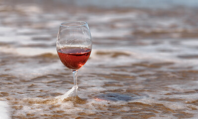 crystal glass with rose wine stands in the sea on the sand, against the background of a sea wave in defocus