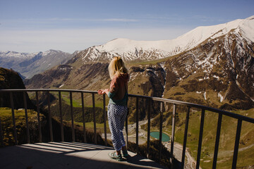 Fototapeta na wymiar A girl with long hair stands against the backdrop of a mountain landscape with snow-capped peaks and a beautiful lake below