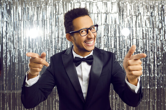 You are chosen for this. Cheerful man makes finger guns gesture directly at camera standing on shiny silver background. Smiling young dark skinned man in black classic suit pointing his fingers at you