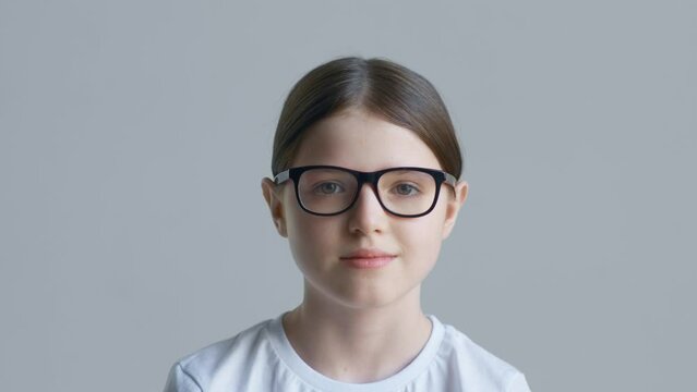 A blonde teenager girl in a white T-shirt puts on and takes off red glasses against the background of a white wall and smiles
