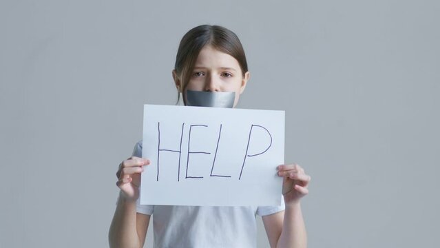 A Teen Blonde Girl In A White T-shirt Stands With Her Mouth Covered With Tape And Holds A Sheet With The Text Help On The Background Of A White Wall