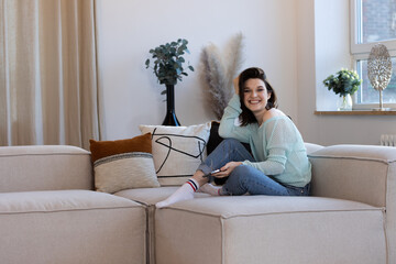 Obraz premium Young woman using her phone sitting at home on her sofa in the living room and smiles