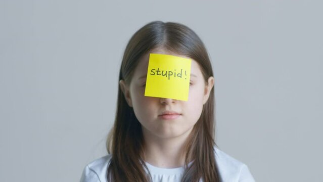 Blonde teenager girl in a white t-shirt hand puts a yellow sticker with the text stupid on her forehead against the background of a white wall