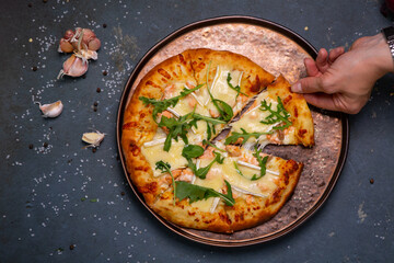 pizza with seafood arugula and cheese lies on an iron plate on a dark background. Mukhskaya hand takes a piece