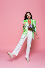 full length of smiling young woman in tie dye blazer holding credit card and flowers on pink.