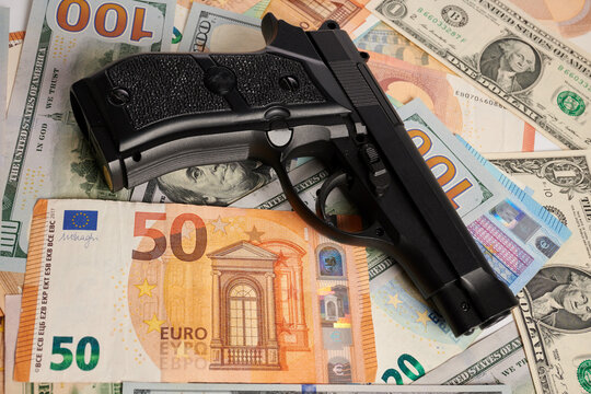 Gun - photo on money bills. Black gun is laying on money bills. Dollar and Euro fiat money under handgun. Weapon protection of business earnings. Pistol over USD and EUR banknotes. Protect your cash.