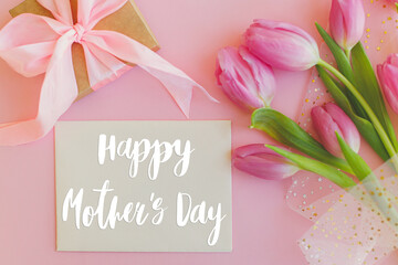 Happy mothers day text on greeting card, pink tulips bouquet and gift box on pink background....