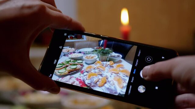 Female Hands Takes a Photo of Food, a Christmas Candle on a Smartphone. Home kitchen. There are a lot of cooked yummy sandwiches, cake on table. Blurred background. Concept holiday, event, New Year.
