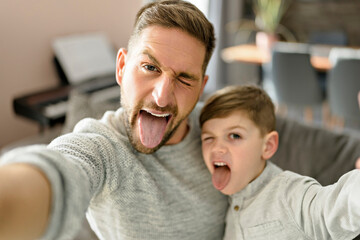 Father and son spending time together on sofa taking selfies and making grimaces - obrazy, fototapety, plakaty
