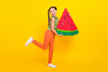 Photo of adorable fooling preteen girl dressed green top holding big watermelon piece walking smiling isolated yellow color background