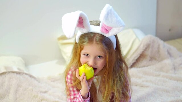 Happy child girl blonde with rabbit ears eats a green apple and laughs. Preparation for the Easter holiday. Easter outfit for a child.