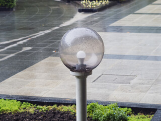 A low, round, wet street lamp with a transparent glass cover in a flower bed against the backdrop...