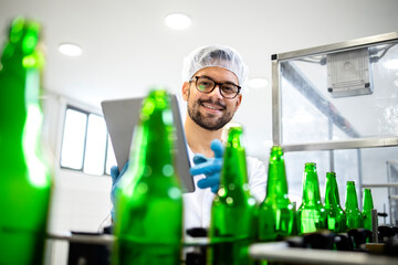 Beer bottles on the conveyor belt and factory worker controlling beverage production in bottling...