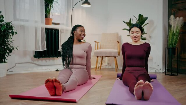 Two diverse women friends warming up their feet and talking, exercising on fitness mats at home