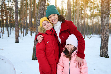 Happy loving mother- beautiful woman gently hugging her adorable kids, boy and girl, smiling with cheerful toothy smile while walking on a snow covered forest path on a winter cool and sunny day