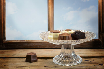 Chocolate pralines on a crystal glass plate on a rustic wooden table in front of a window, copy...